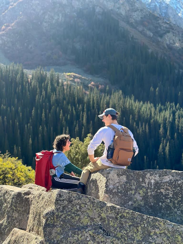 Two students sit on rocks on a sunny day with a beautiful forest in the background.