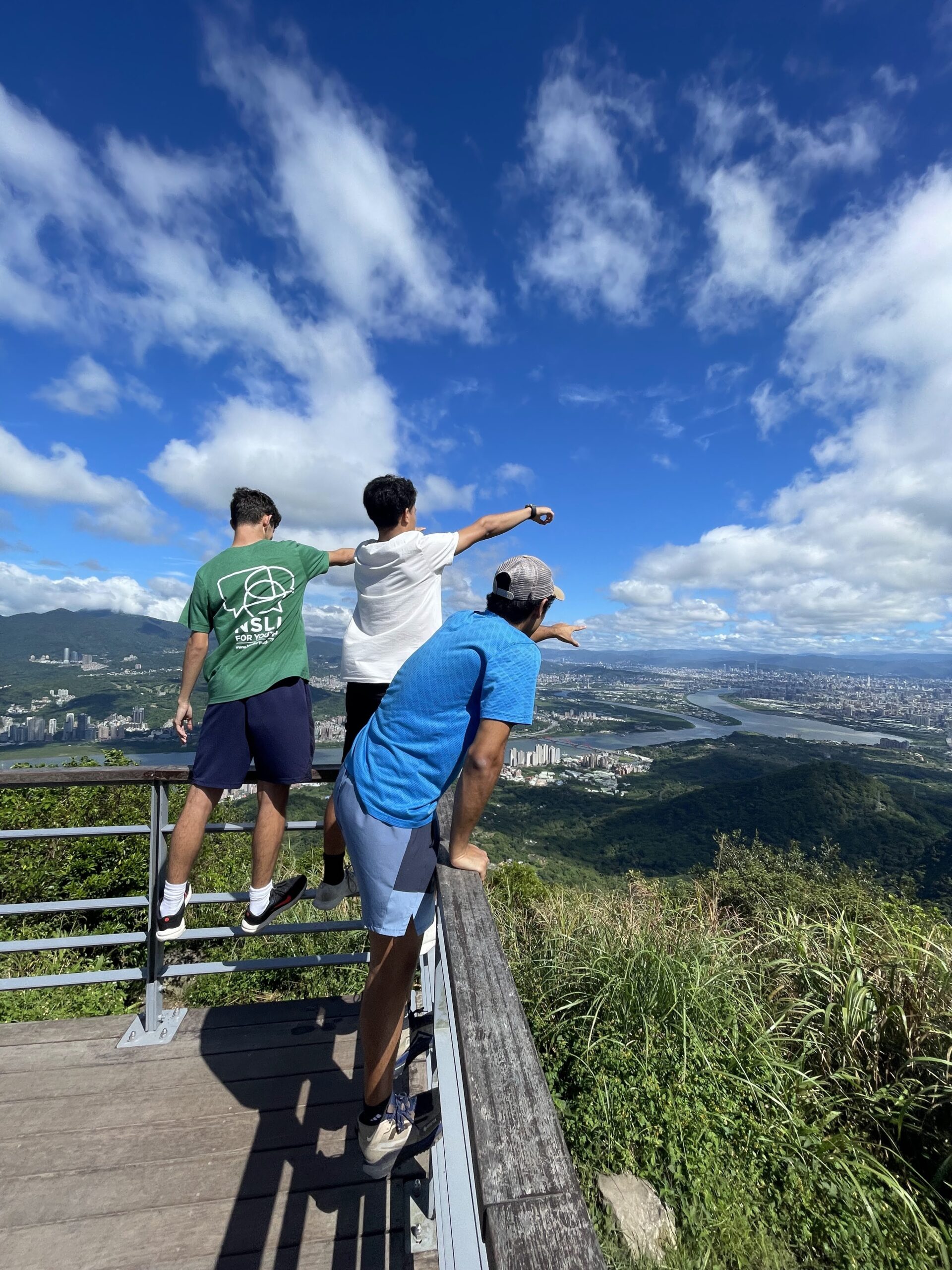 Three young men look from a scenic overlook at sunny Taipei below.