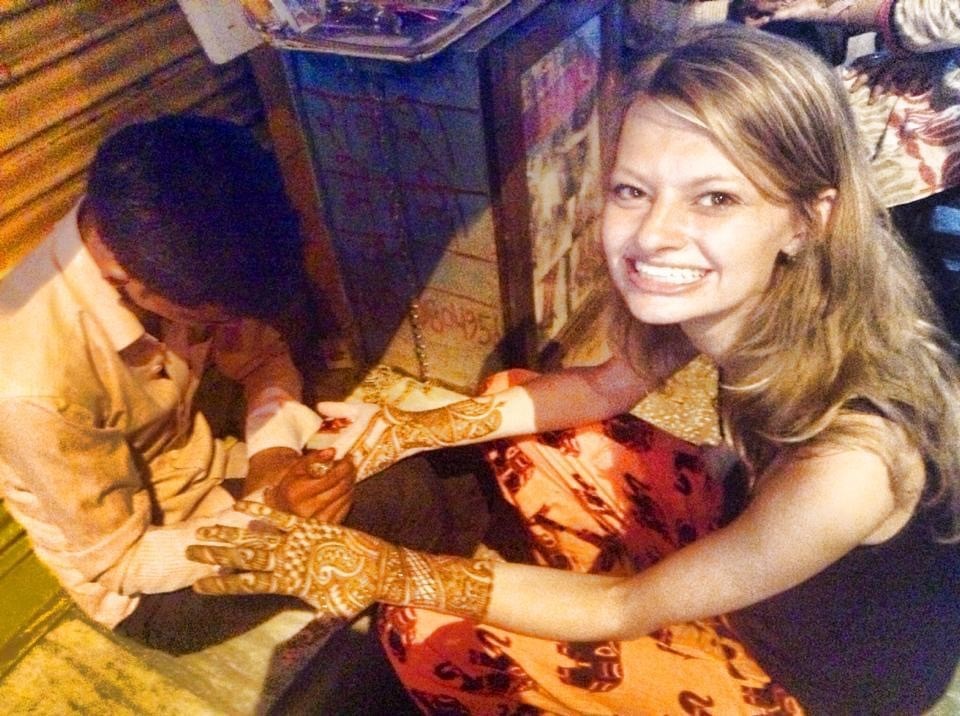 Taylor wearing orange pants and a black top getting mehendi (henna) done on her hands. The artist is sitting in front of her wearing a white shirt.