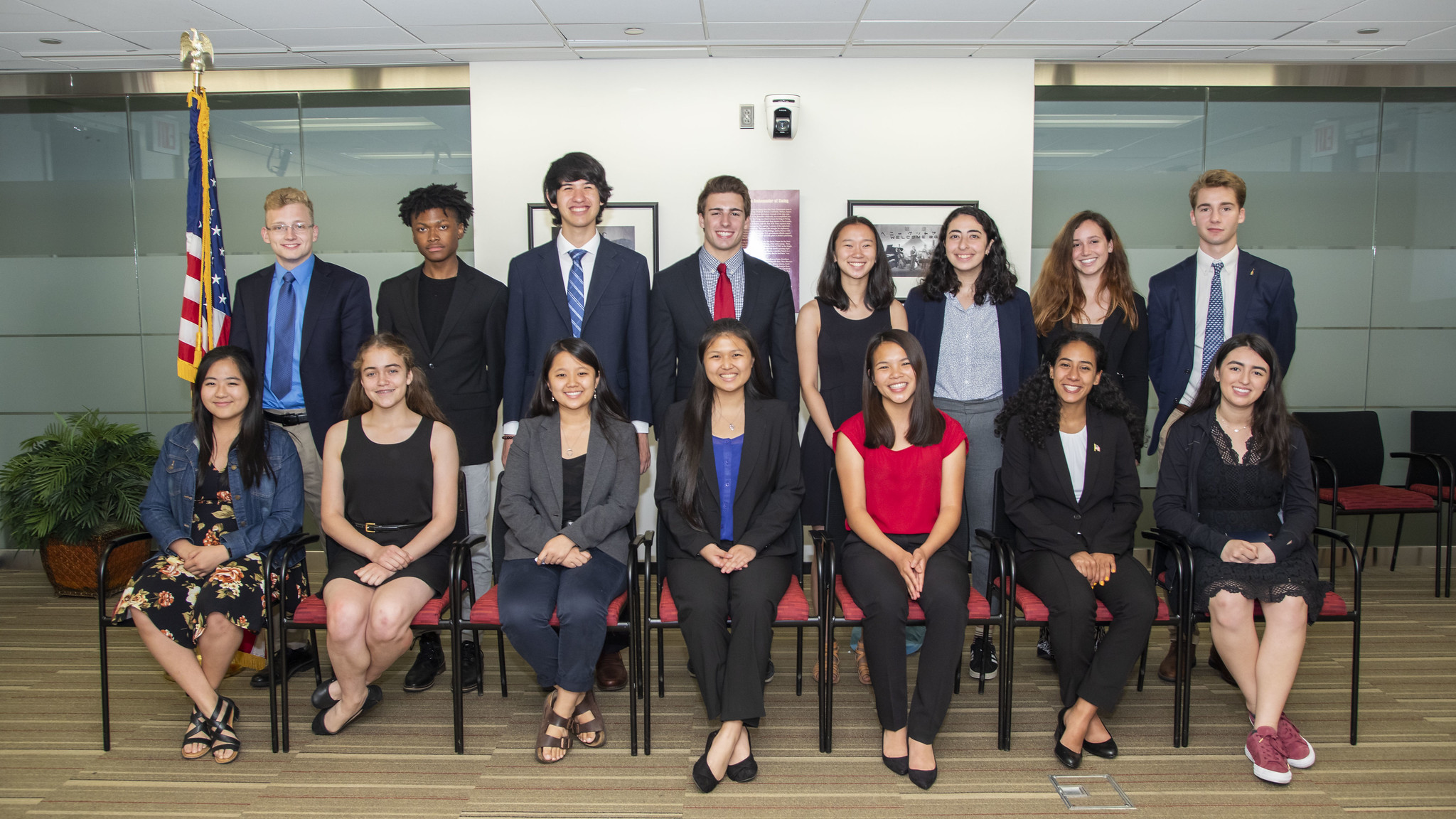A picture of 15 students in an office building. Eight students are standing in the back row and seven students are seated in the front row. They are all wearing business casual clothing. There is an American flag to the left side of the students.